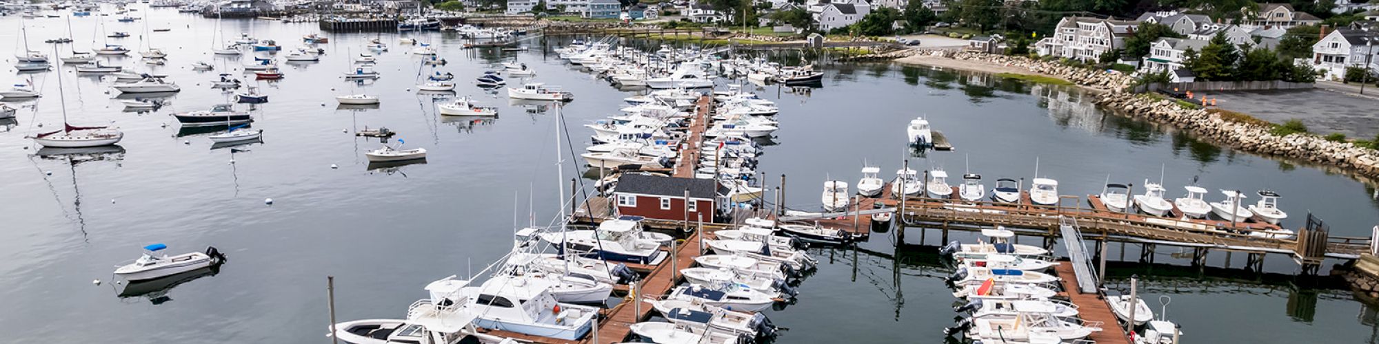 An aerial view of a marina filled with boats docked along wooden piers, with a coastal town and numerous other boats in the background.