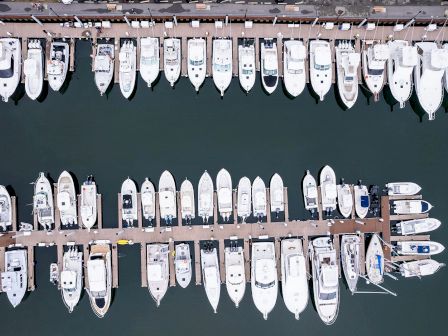 An aerial view shows two rows of docked boats aligned parallel to each other with a walkway in between on a calm body of water.