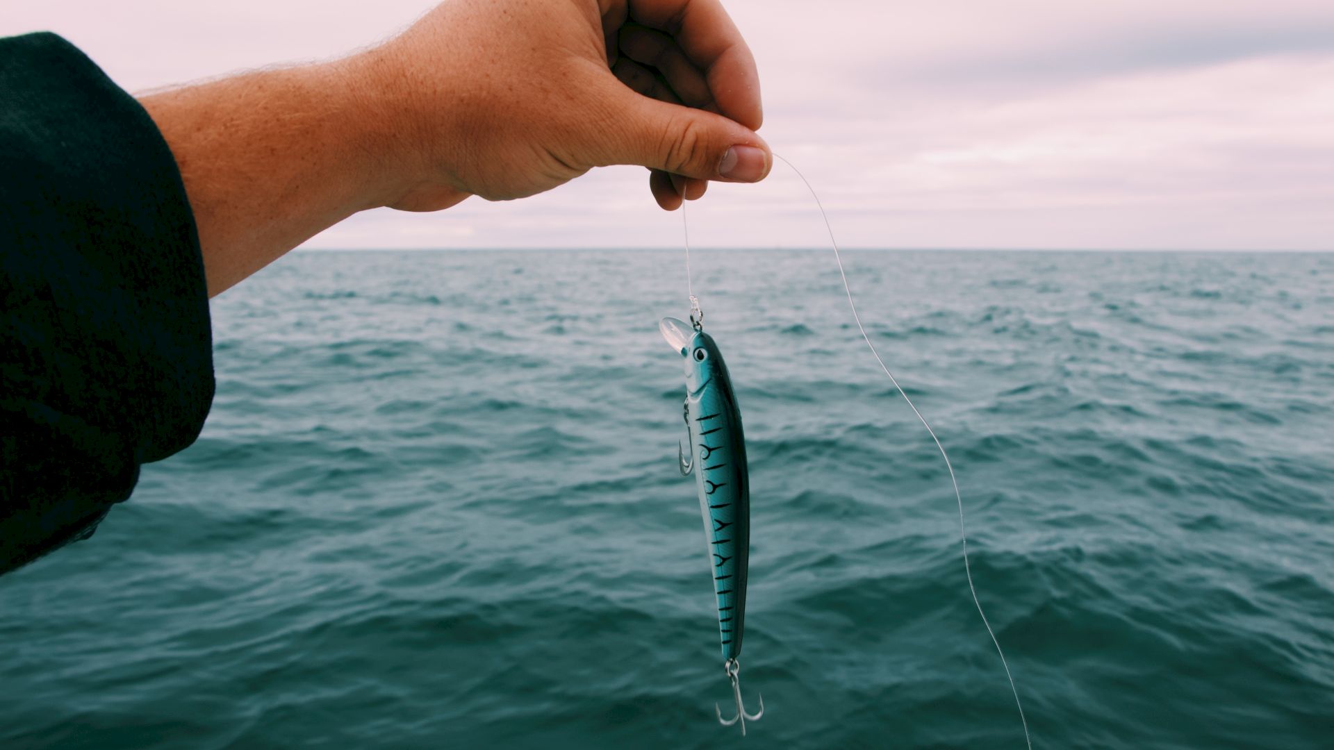 A hand holding a fishing lure above the ocean with cloudy skies in the background.
