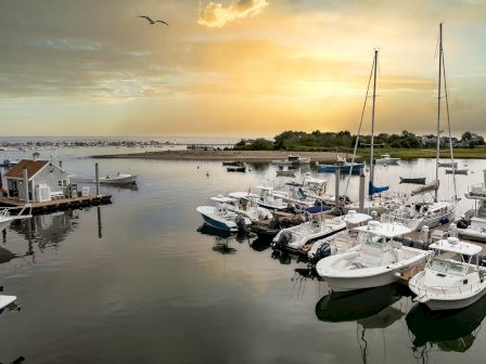 A tranquil marina at sunset with numerous boats docked, calm waters reflecting the sky, and birds flying overhead, exuding peace and serenity.