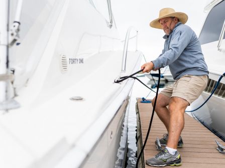 A person is tying up a boat at a dock while wearing a hat, shorts, and a long-sleeve shirt, with another boat visible in the background.