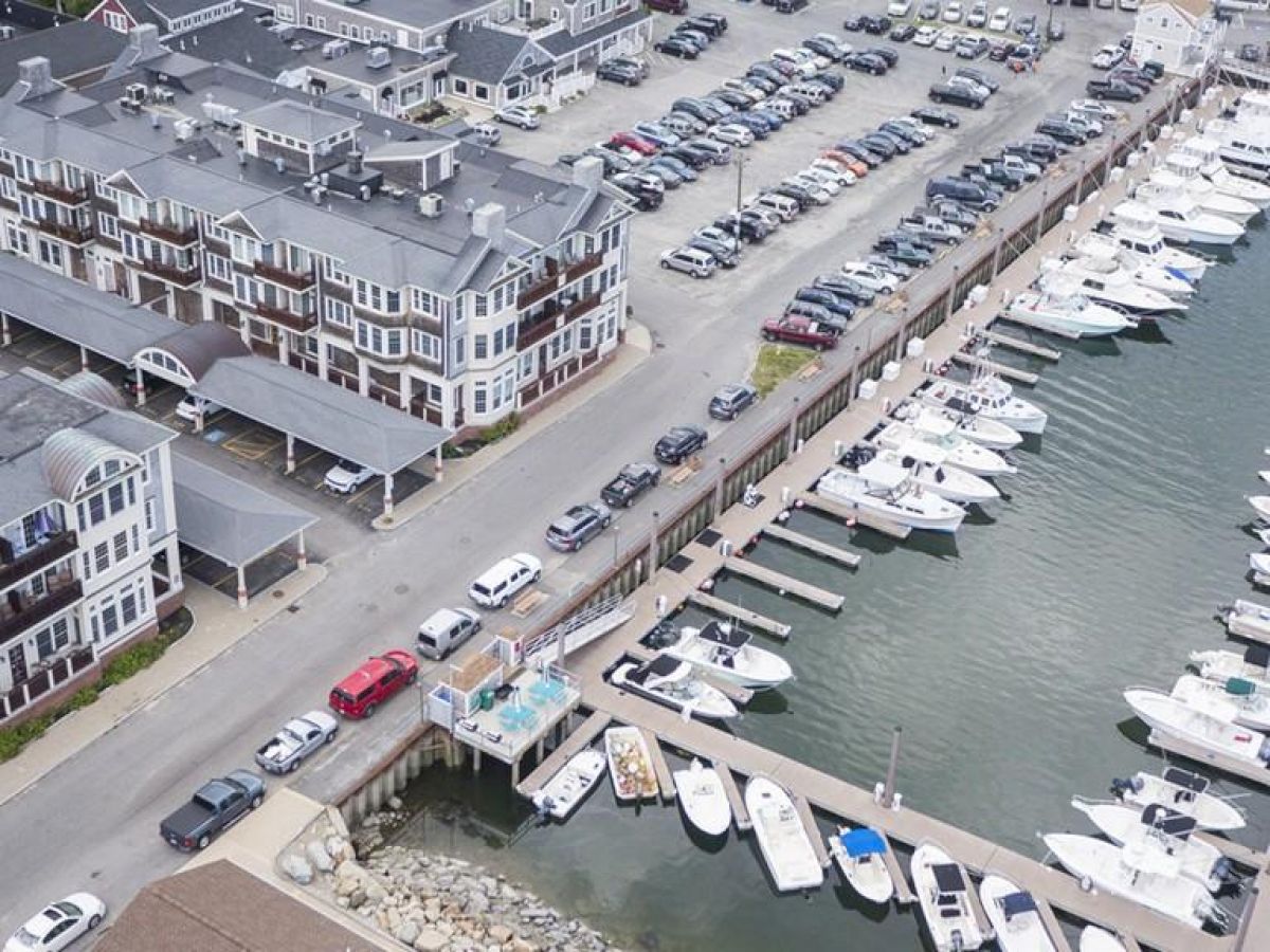 The image shows a marina with parked boats, adjacent to a coastal town with buildings and parked cars, under a slightly overcast sky.