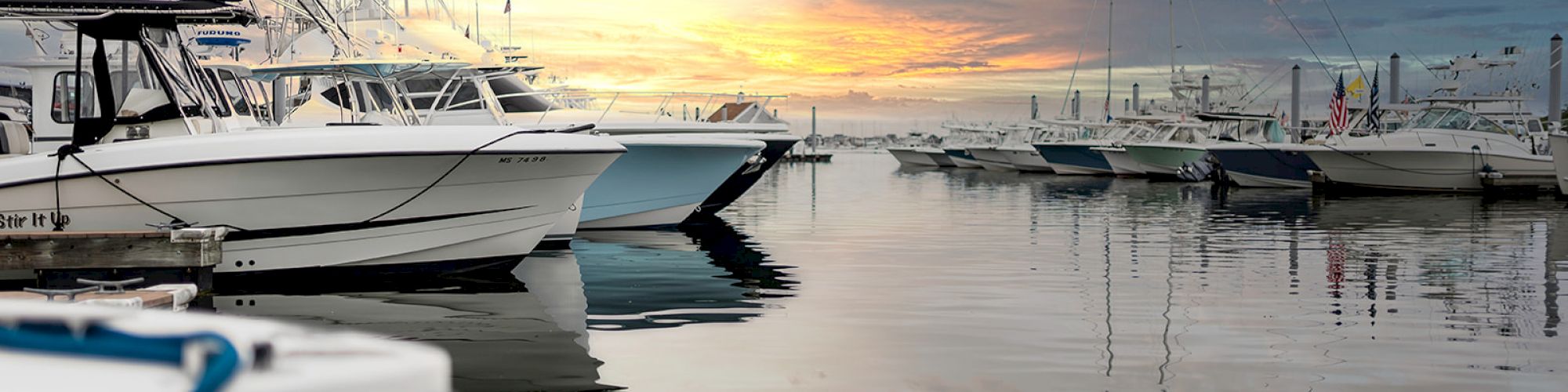 Several boats are docked at a marina during sunset, with calm water reflecting the sky's colors and some clouds present.